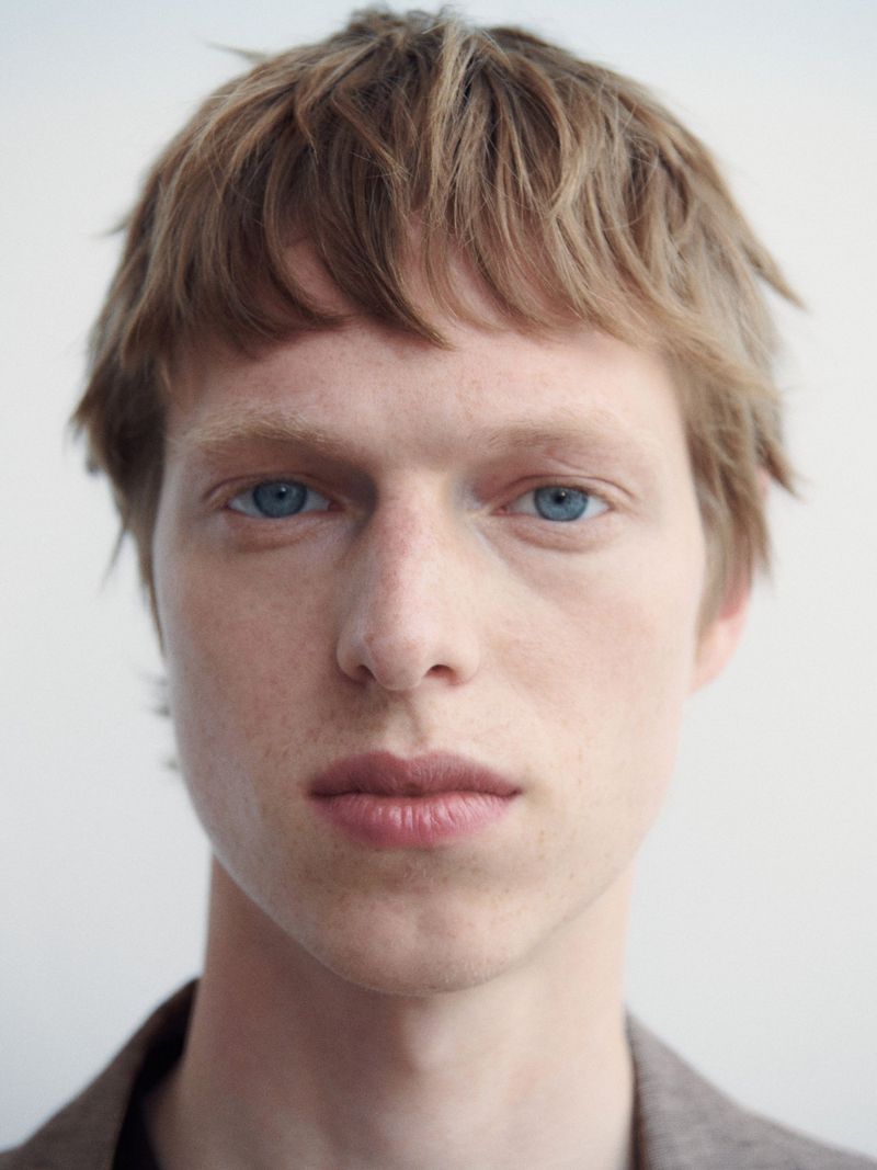 Close-up portrait of a young adult male with light brown hair and blue eyes, wearing a neutral-toned jacket against a plain white background.