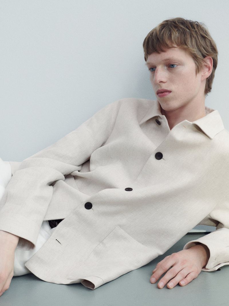 A young adult man with light brown hair and a linen jacket poses in a studio with a light blue background. He wears a casual outfit.