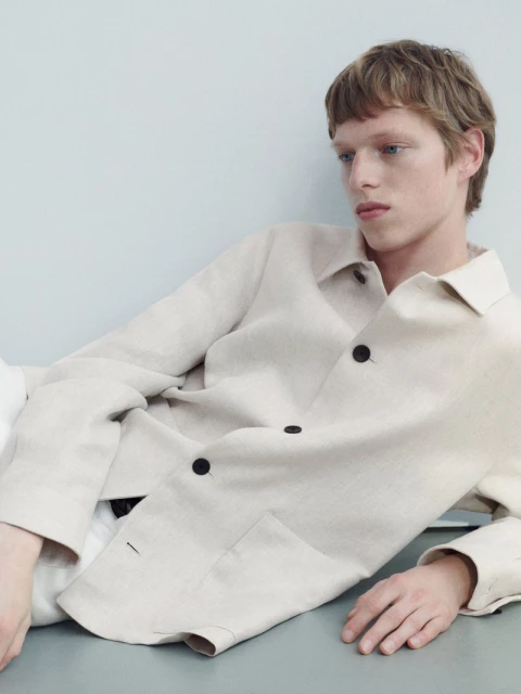 A young adult man with light brown hair and a linen jacket poses in a studio with a light blue background. He wears a casual outfit.