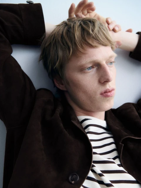 A young, light-skinned man with blond hair poses indoors with a brown jacket and striped shirt, lying down with his hands behind his head.