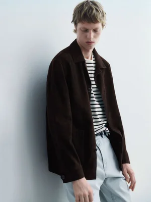 A young male model leaning against a plain wall, wearing a dark brown jacket, striped t-shirt, and light denim jeans.