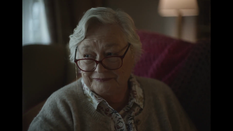 Close-up shot of a smiling elderly woman with glasses and short white hair wearing a patterned blouse and cardigan in an indoor setting.