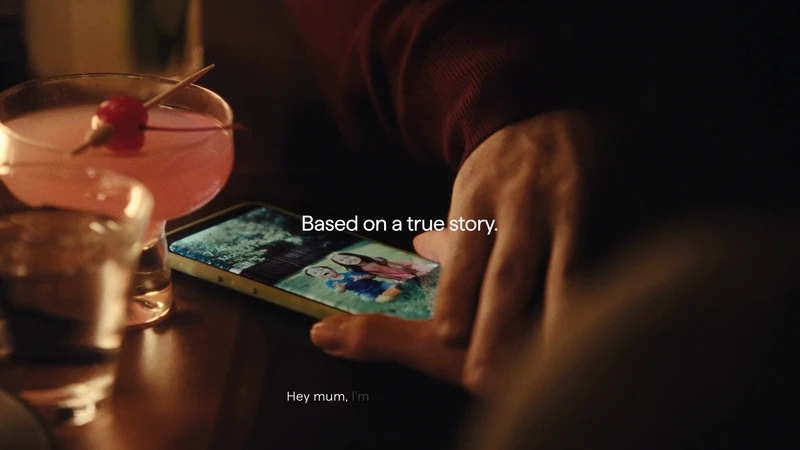 Close-up shot of a person holding a smartphone displaying a photo of two children and an adult, next to a pink cocktail.