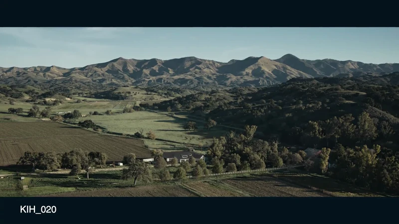 Aerial shot of rolling hills and valleys, with green fields and a home nestled among trees in the foreground, under a clear blue sky.