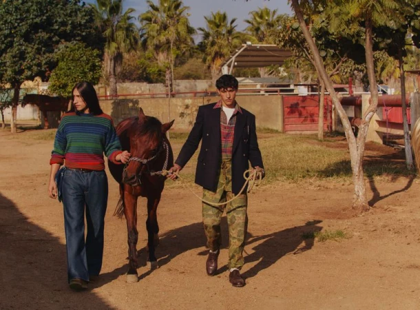 Two men walking a brown horse along a sunlit dusty path past palm trees and stables for Mr Porter
