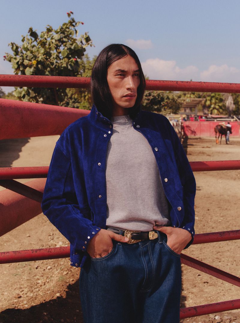 Adult male in jeans and blue jacket poses outdoors with rural background on sunny day looking off camera in a fashion style shot.