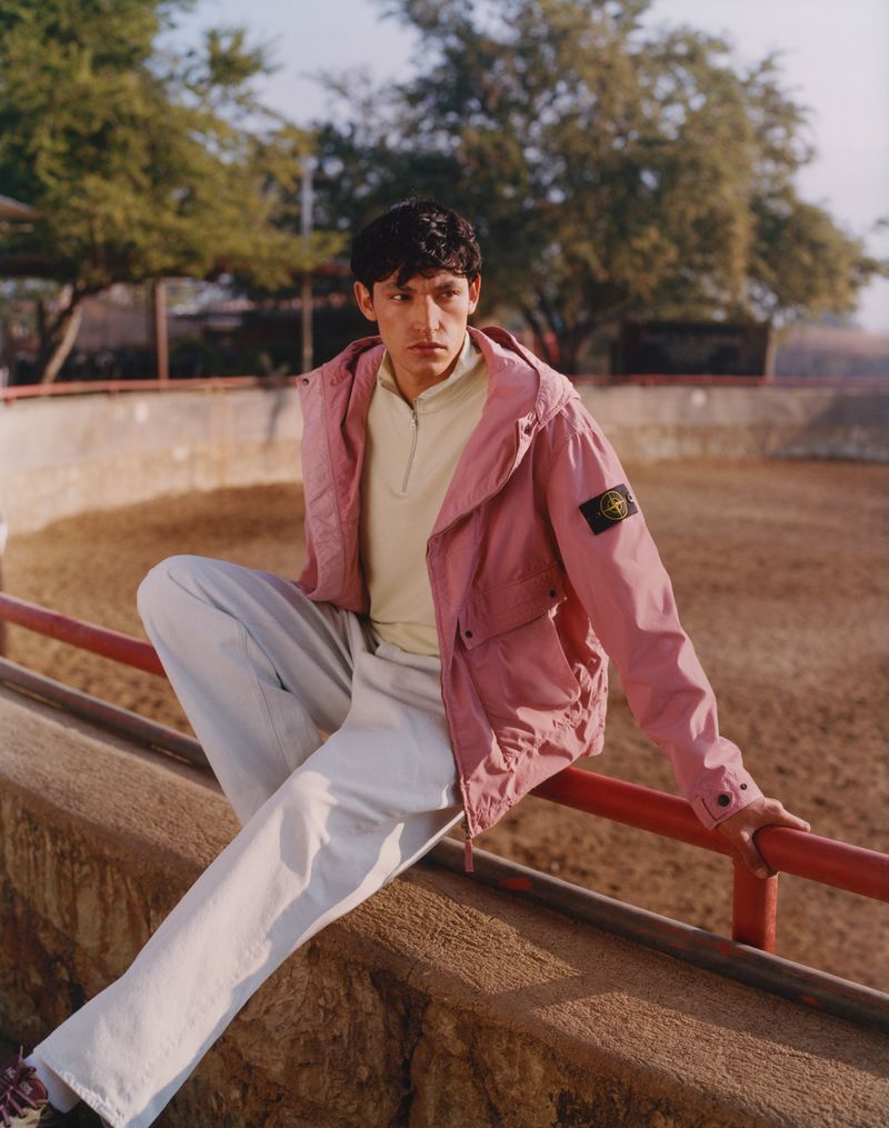 An adult male model poses outdoors wearing a pale yellow polo shirt, loose-fitting white pants, and a pink hooded jacket.