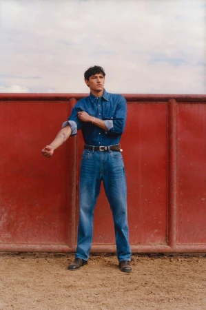 Man wearing a blue denim shirt and jeans rolling up his sleeve in front of a red wall