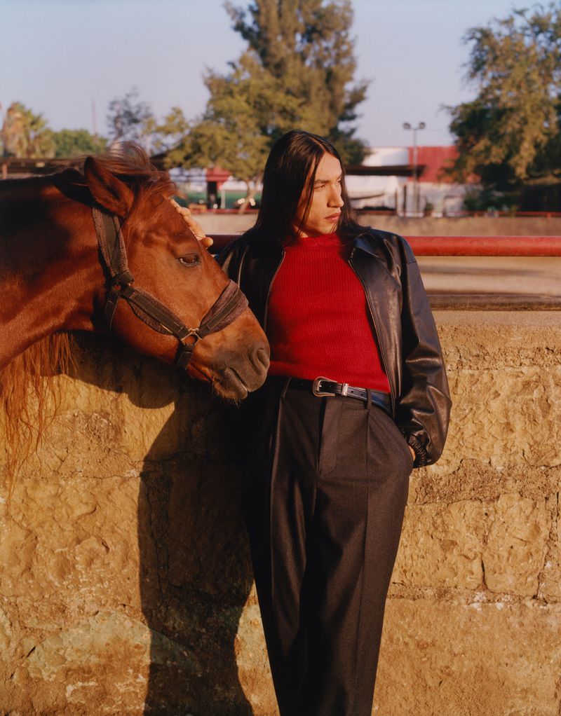 Man and horse stand together outside in a ranch setting with a tan wall, trees, a red sweater and black leather jacket and pants.