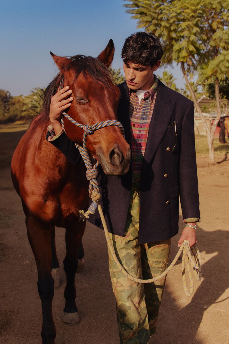 Man and brown horse stand outdoors. Man in plaid shirt and blazer holds horse's lead. Trees and structures are in the background.