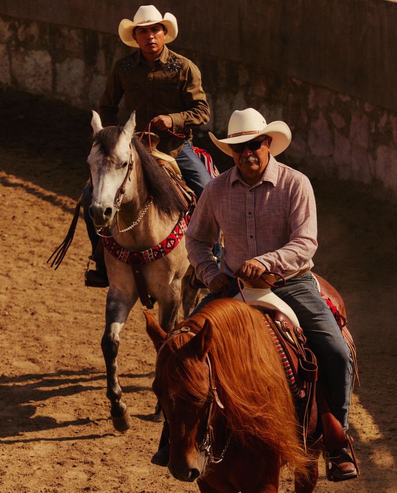 Two cowboys riding horses outdoors in arena, wearing hats and western attire, brown and grey horses, dirt ground.