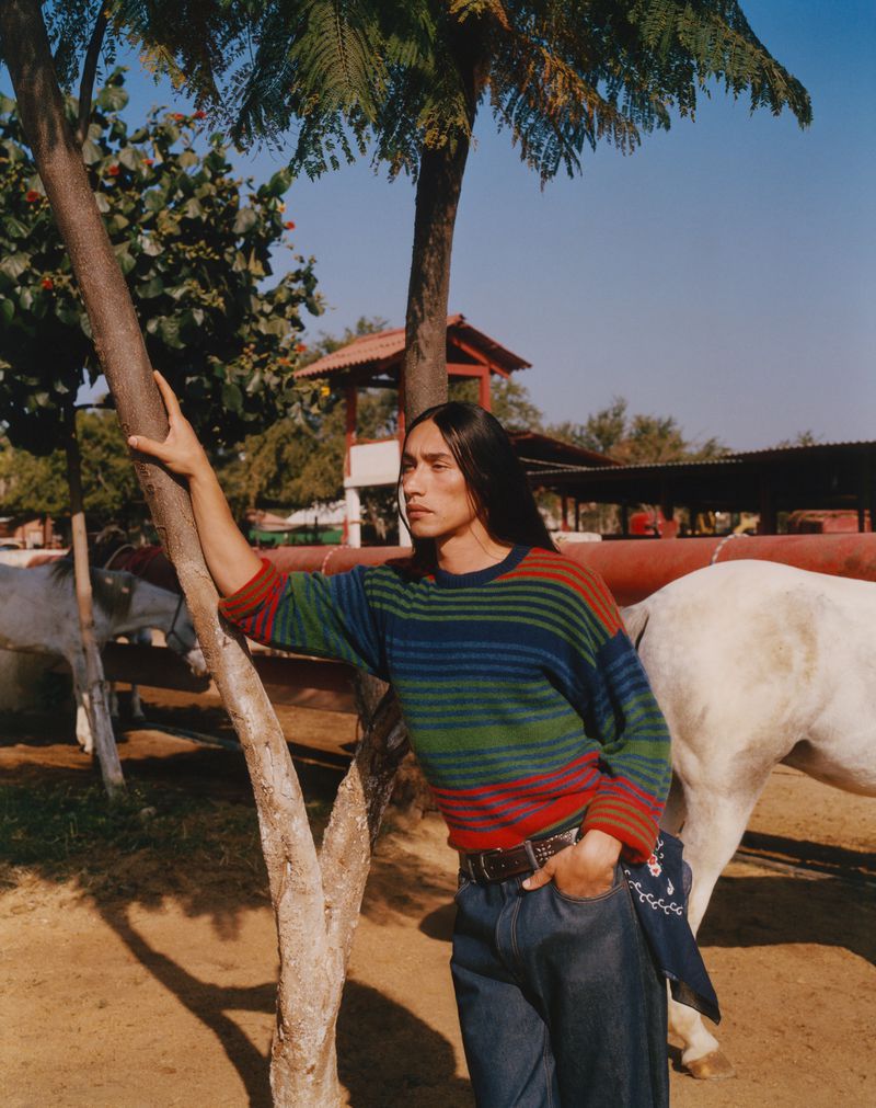 An adult man with long dark hair poses in an outdoor setting, wearing a colorful striped sweater, jeans, and a bandana, next to a horse.