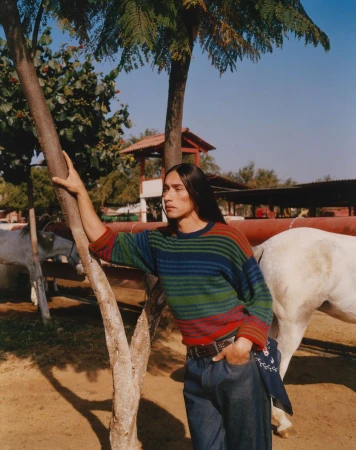 An adult man with long dark hair poses in an outdoor setting, wearing a colorful striped sweater, jeans, and a bandana, next to a horse.