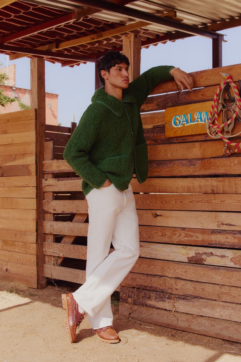 Man in green sherpa jacket leaning against a wooden fence outdoors in sunlight, wearing white pants and brown leather dress shoes.