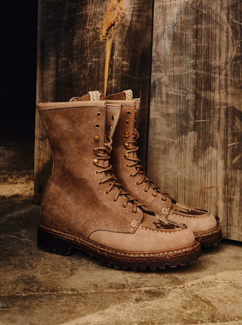 A pair of ankle-high, tan-colored, lace-up boots with dark brown soles are set against a wooden backdrop, suggesting a rustic fashion style.