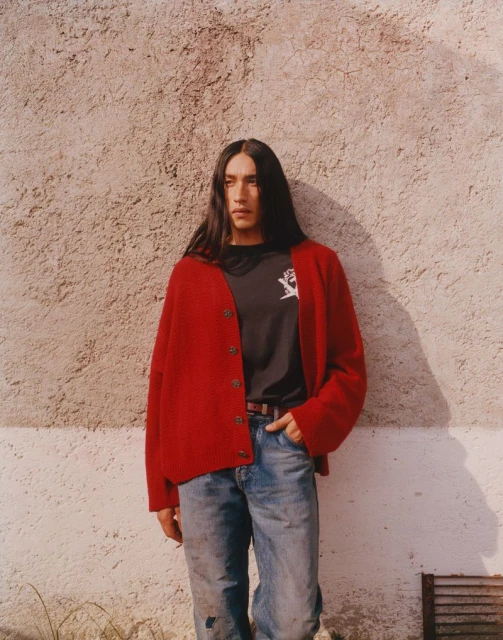 Man in red cardigan and jeans poses against textured wall; long dark hair; casual fashion; daytime; outdoor setting, natural light, male model.