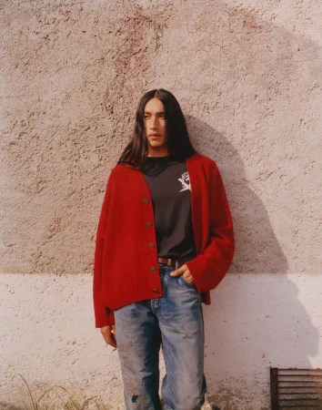 Man in red cardigan and jeans poses against textured wall; long dark hair; casual fashion; daytime; outdoor setting, natural light, male model.