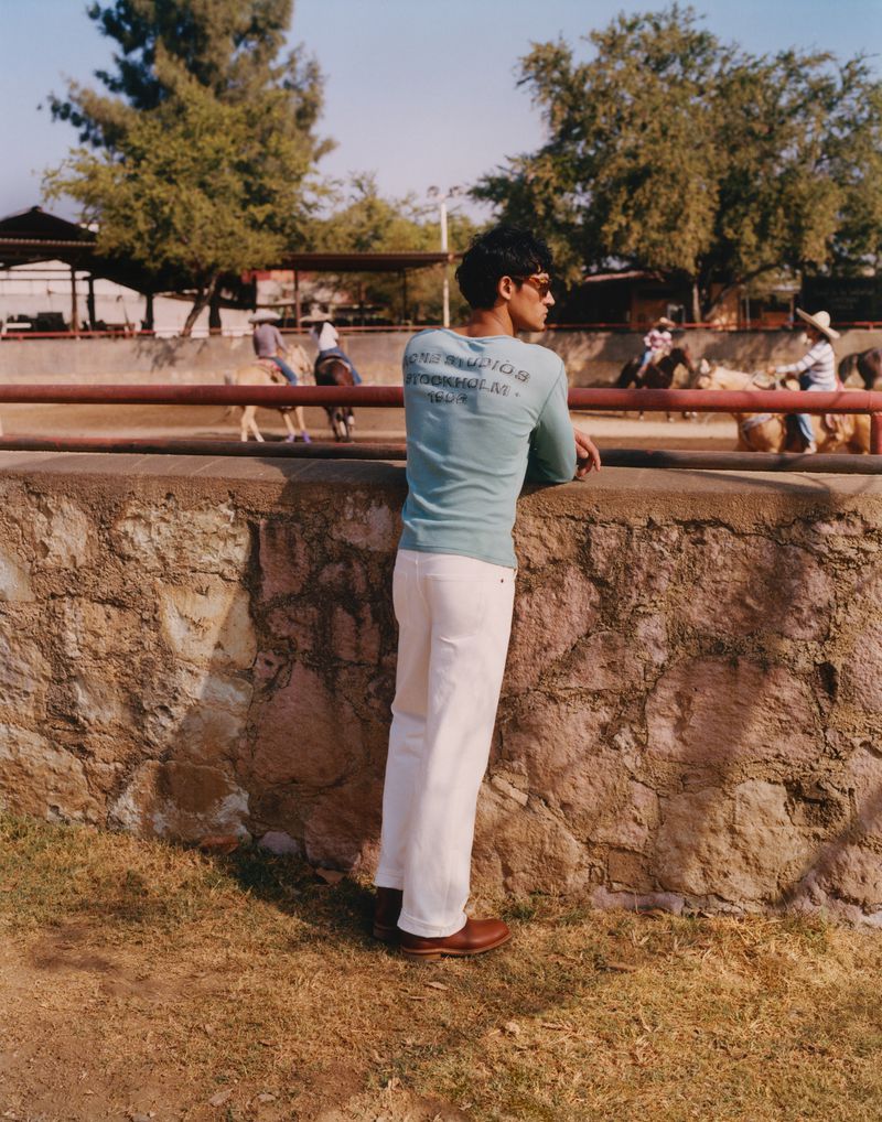 Adult male in a light blue shirt and white pants leaning against a stone wall; horse riding in the background on a sunny day.