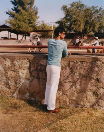 Adult male in a light blue shirt and white pants leaning against a stone wall; horse riding in the background on a sunny day.