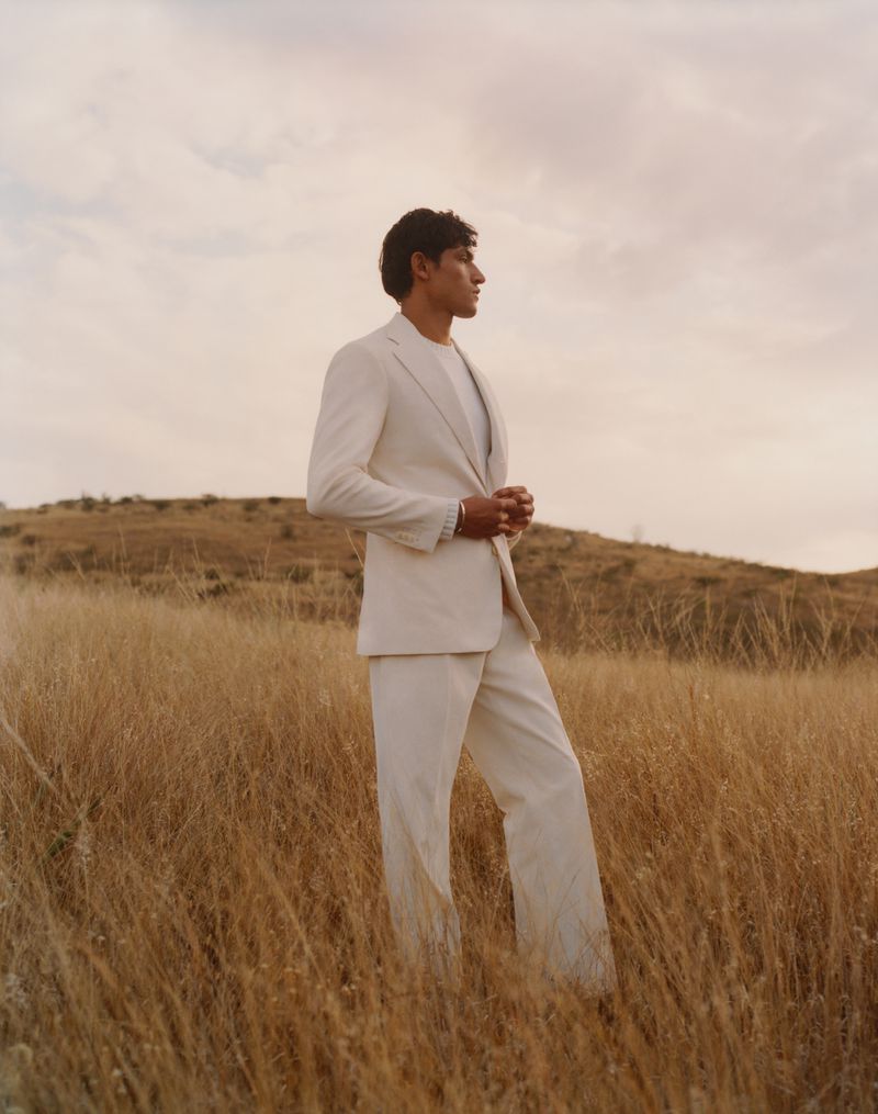 Man wearing a cream suit standing in a dry grassy field under an overcast sky.