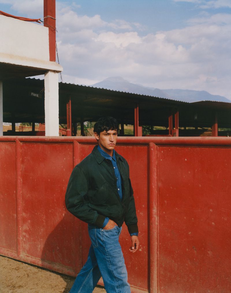 Man in denim shirt and jeans walks in front of the red wall of a bullfighting ring with mountains and cloudy blue sky in background.