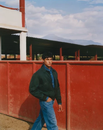 Man in denim shirt and jeans walks in front of the red wall of a bullfighting ring with mountains and cloudy blue sky in background.