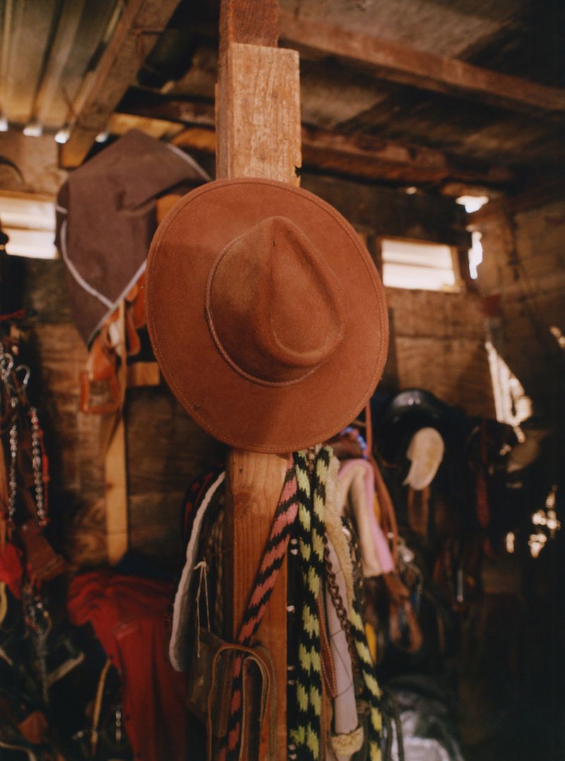 Image shows a brown cowboy hat hanging on a wooden post indoors, with other hats, and garments in the background, creating a rustic scene.