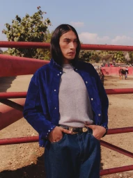 Man with long dark hair wearing a blue jacket standing outdoors against a red fence for Mr Porter photographed by Vivek Vadoliya