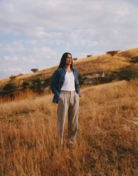 A man with long dark hair stands in a dry grassy meadow under a cloudy sky, wearing a blue shirt over a white t-shirt and grey trousers.