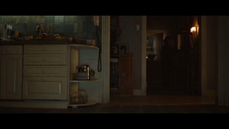 Dimly lit kitchen with white cabinetry, various objects on countertops, and a glimpse into a darker adjacent room.