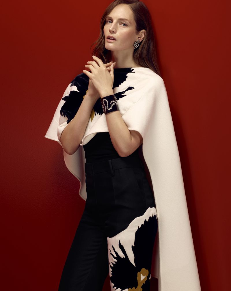 A female fashion model poses against a red background in a stylish black-and-white outfit and jewellery, captured in a studio shot.