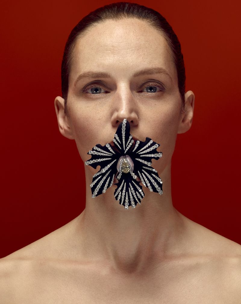 Striking beauty shot of a woman with fair skin and an orchid covering her mouth against a red background in soft, diffused lighting.