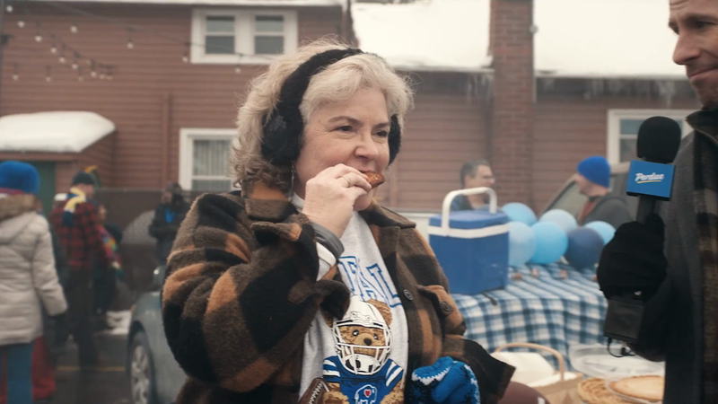 Woman with earmuffs eating food outdoors near reporter with microphone, balloons, and a building with icicles in winter.