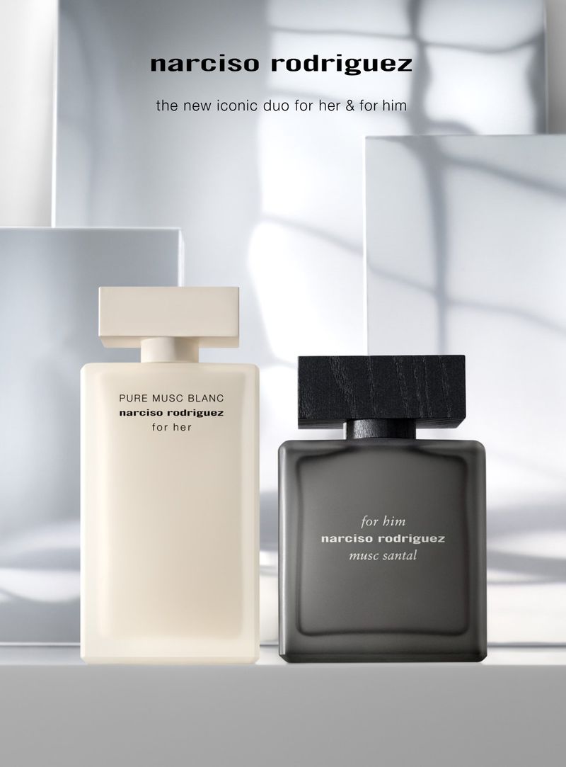 Two Narciso Rodriguez fragrance bottles, one white and one dark, stand on a white surface against an out-of-focus light background.