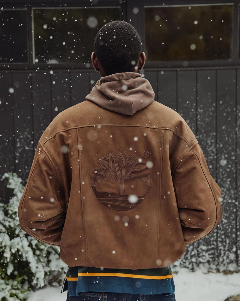 Man wearing brown Timberland jacket and a hoodie stands in snowy weather with a dark fence in the background. Medium shot, outdoors.