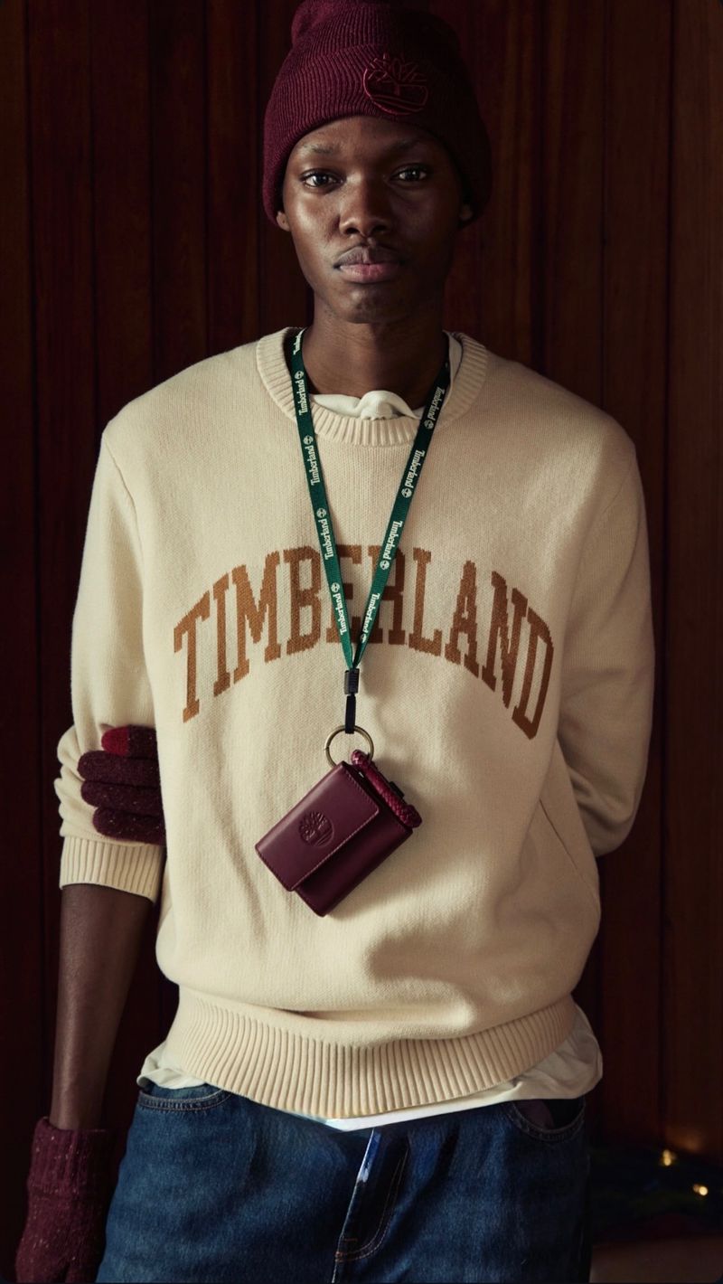 Close-up of an adult male fashion model wearing a beige Timberland sweatshirt, jeans, and a burgundy beanie in a wood-panelled studio.