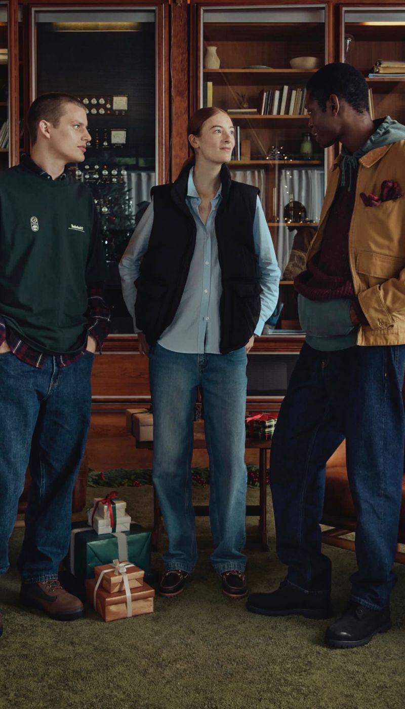 Three people—two men and one woman—stand indoors near gifts and a bookcase, wearing casual clothing, in a medium portrait shot.