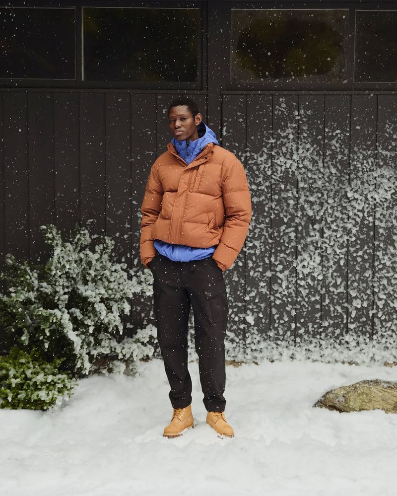 Man in an orange puffer jacket and black pants standing in a snowy outdoor environment against a dark grey building on a winter day.