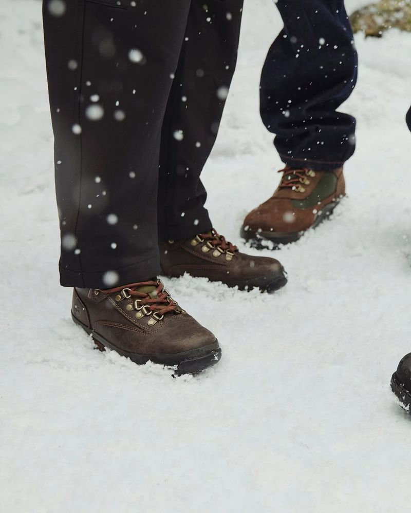 Three people stand in the snow wearing winter boots and dark colored pants in an outdoor wintery environment with falling snow.