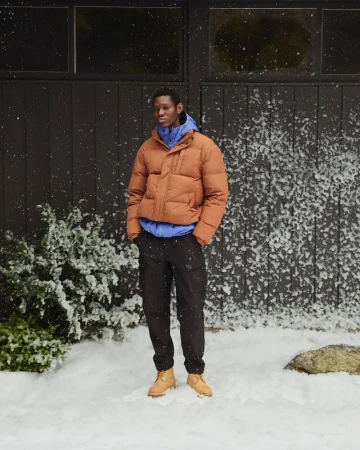 Man wearing a brown puffer jacket, blue hoodie, dark trousers, and tan boots standing outdoors in snowy weather.