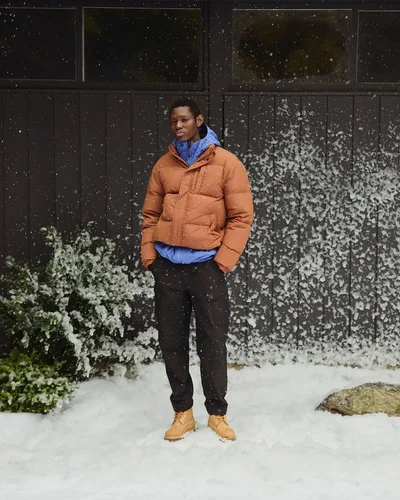 Man wearing a brown puffer jacket, blue hoodie, dark trousers, and tan boots standing outdoors in snowy weather.