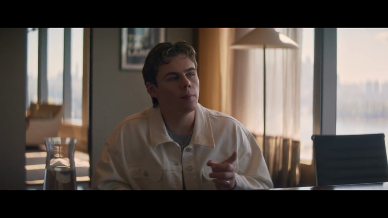 Young man sits at a table indoors with a carafe, buildings visible through the window. He wears a cream jacket and silver chain.
