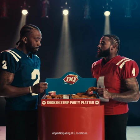 Two football players pose with a DQ chicken strip platter in a promotional shot, spotlighted against a dark backdrop, promoting a party.