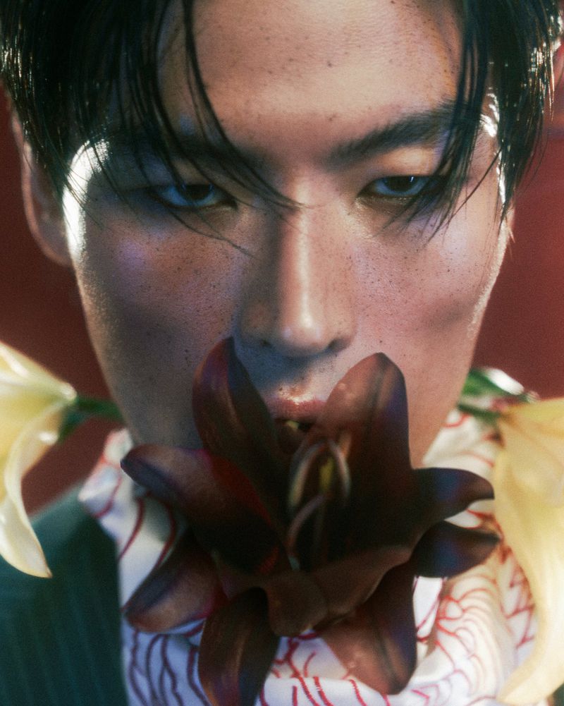 Close-up of a dark-haired person with flowers near their mouth in a beauty shot, wearing patterned fabric and a dark jacket.