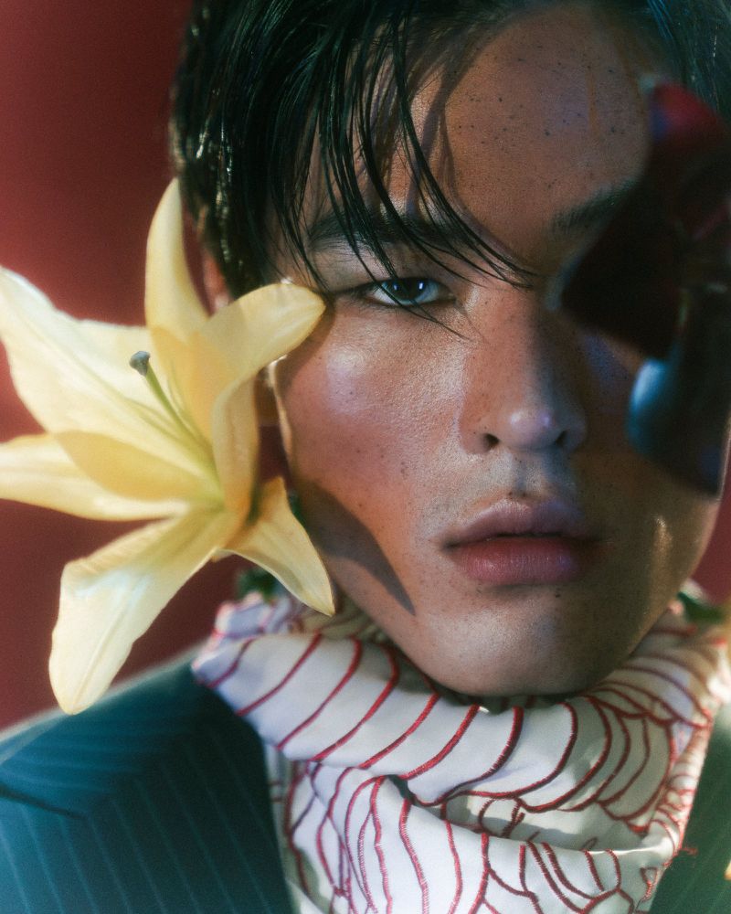 Close-up of an Asian male model with flower near his face, wearing striped suit and patterned scarf against a red backdrop.