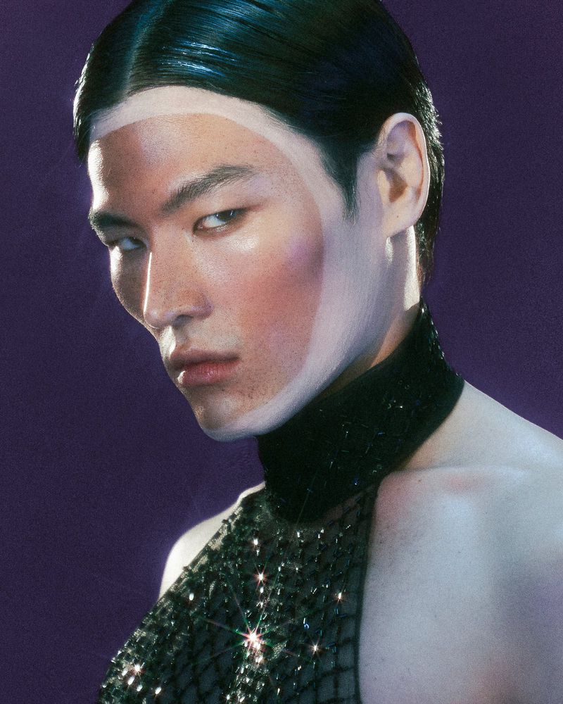 Close-up portrait of an Asian male model with dramatic lighting, wearing a black sparkling halter top against a deep purple backdrop.