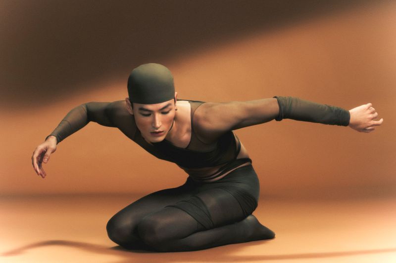 Adult male kneeling in a forward reaching pose with arms outstretched wearing a black swim cap and sheer top in a studio setting.