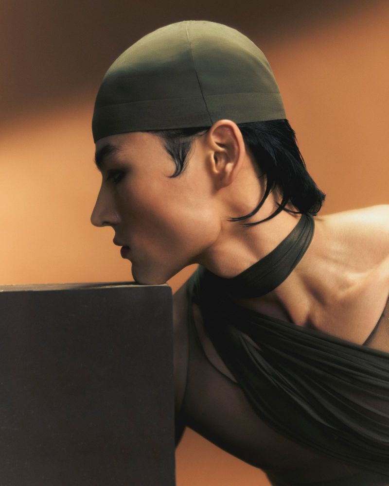 Profile view of an adult in a fashionable olive-green head covering, draped in a sheer dark fabric, posed against a warm brown backdrop.