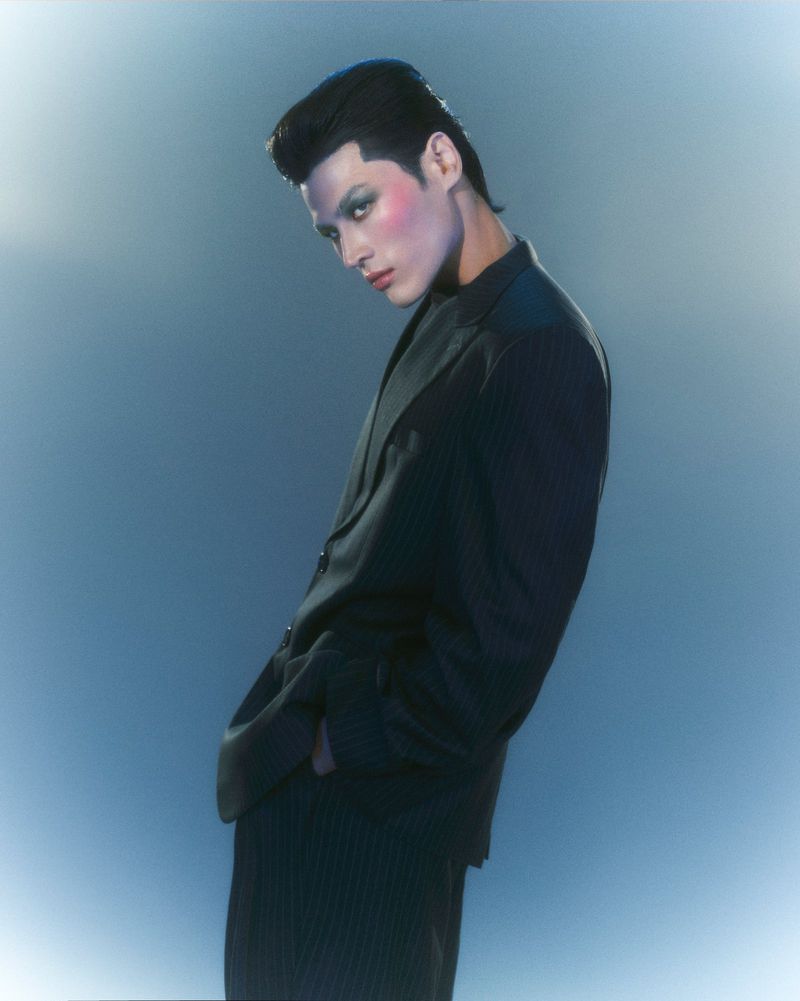 Portrait of a young adult man in a black suit with retro-style hair and makeup, posing against a graduated blue background in a studio setting.