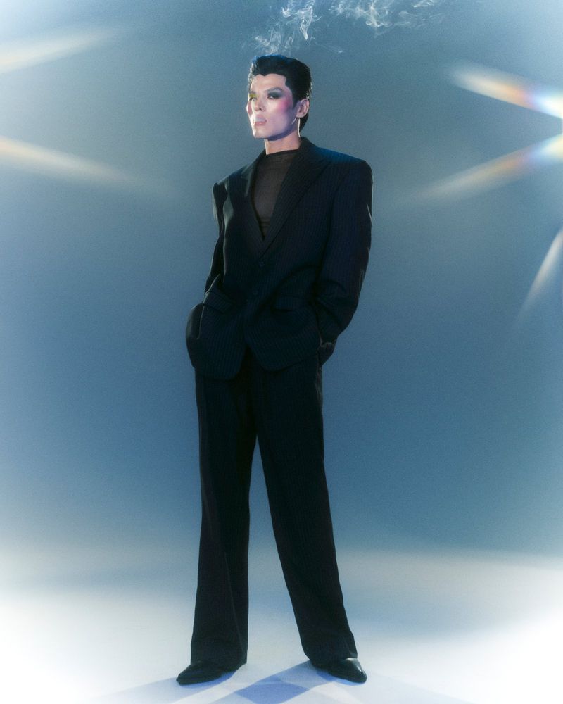 Stylish adult posing in a black suit against a blue background with smoke detail, standing in full shot in a fashion portrait.